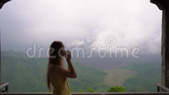一名年轻女子在山上的一家咖啡馆喝茶或咖啡观看巴图山和视频的预览图