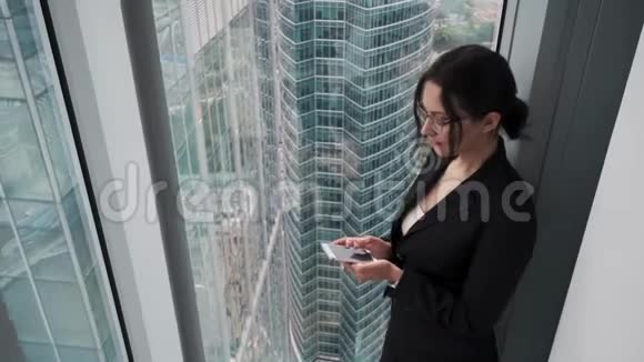 穿着商务服的年轻女性站在摩天大楼的高楼层上时使用智能手机视频的预览图