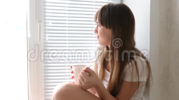 小女孩早上在窗边喝茶美丽的女孩看着窗外手里拿着一杯茶视频的预览图