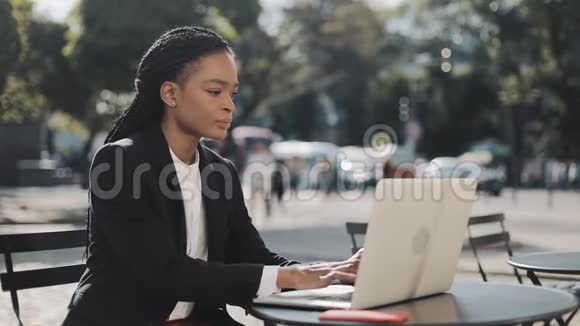 一位非洲裔美国青年妇女坐在夏季露台的咖啡馆里在笔记本电脑上工作商务工作视频的预览图