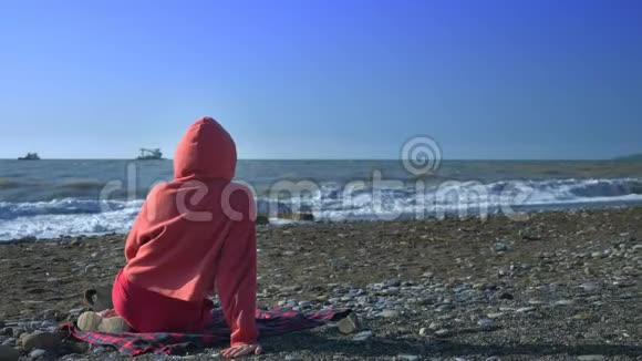 后景有人穿着红色运动衫和引擎盖在暴风雨中坐在海滩上看着大海视频的预览图