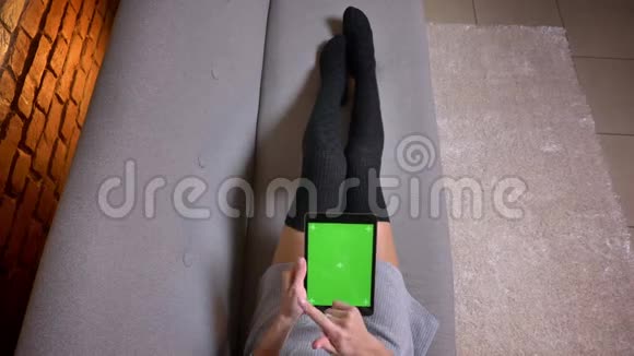 室内沙发上穿着可爱袜子的年轻女孩腿的特写镜头嫩嫩的女性拿着绿色屏幕的平板电脑视频的预览图