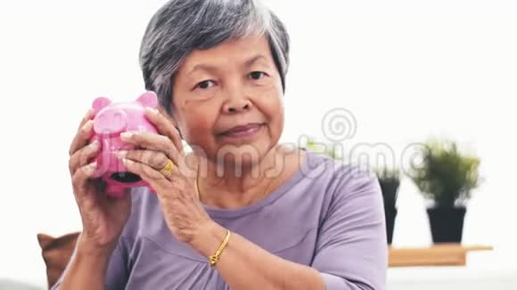 一位兴高采烈的亚洲老太太对着镜头微笑着摇着存钱罐成年女子举着储蓄罐存钱的画像印视频的预览图