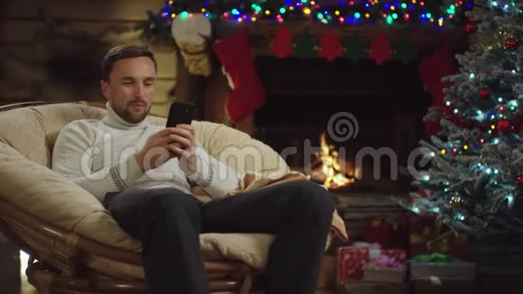 尊敬的成年男子在圣诞夜用装饰好的树在智能手机上发短信视频的预览图