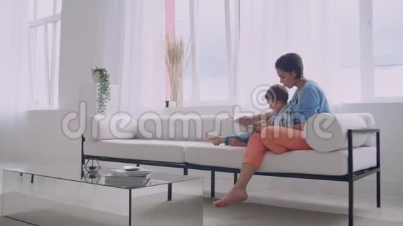 一位年轻的母亲带着一个孩子读一本书坐在客厅里明亮的白色屋子里视频的预览图