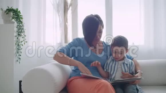 一位年轻的母亲带着一个孩子读一本书坐在客厅里明亮的白色屋子里视频的预览图
