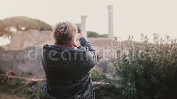 在意大利奥斯蒂亚带着智能手机拍摄一张古老遗址的照片的快乐高颜值白人女性度假后景视频的预览图
