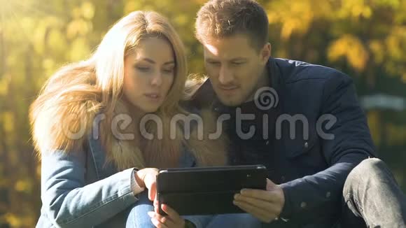 使用平板电脑的快乐夫妻坐在秋天的公园里购物或选择旅游视频的预览图