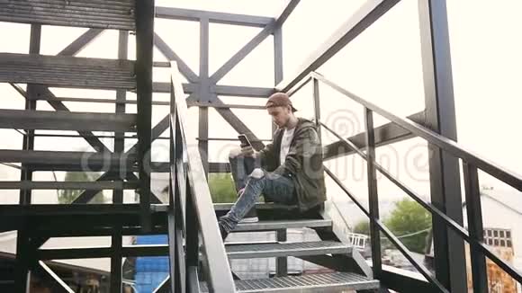 英俊的时髦男人用智能手机坐在工业大楼的铁楼梯上很漂亮的年轻人视频的预览图