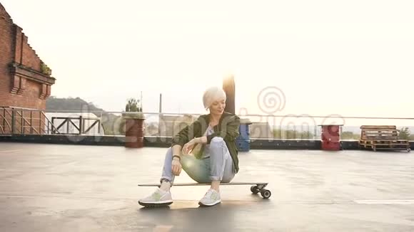 可爱的少女坐在屋顶现代建筑的滑板上年轻的金发女人坐在长板上视频的预览图