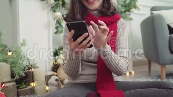 快乐的年轻亚洲女人在圣诞节期间用智能手机在家里的客厅里查看社交媒体视频的预览图