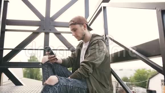 年轻英俊的嬉皮士在日落时分在工业大厦的黑色铁楼梯上使用智能手机年轻人视频的预览图
