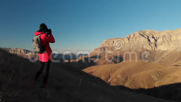 在日落时分一位带着背包和照相机的女孩旅行者在山上拍照女摄影师视频的预览图