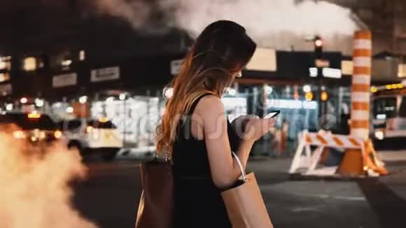 年轻漂亮的女人带着购物袋站在美国纽约的交通道路附近使用智能手机视频的预览图