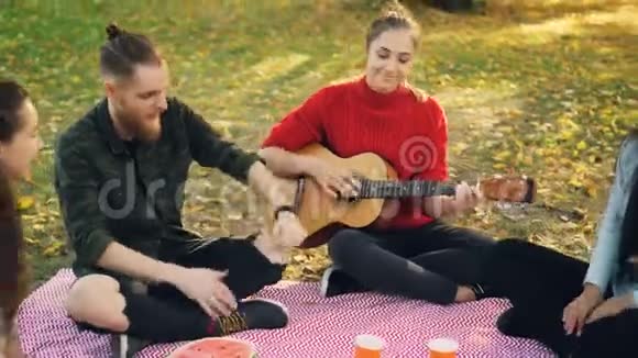 有创造力的年轻女士正在弹吉他而她的男女朋友正坐在格子布上唱歌跳舞视频的预览图