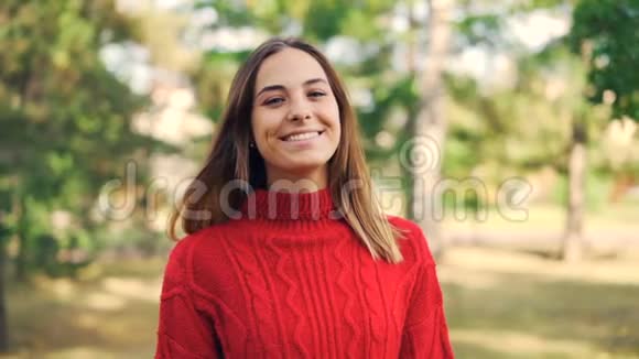 美丽的年轻女子穿着鲜艳的红色毛衣微笑着站在公园里看着公园的慢镜头视频的预览图