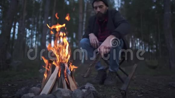 坐在靠近火堆的椅子上试着烧一根木头的小胡子森林里的火堆旁视频的预览图
