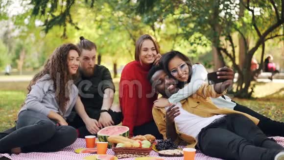快乐的年轻人慢动作时髦的人用智能手机在野餐时自拍男人和女人摆姿势视频的预览图