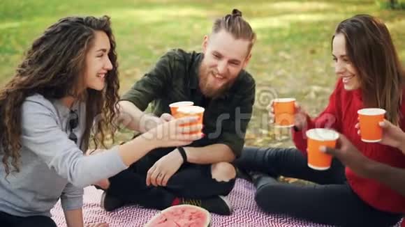 在公园野餐时快乐的女孩和男孩的朋友会慢慢地碰杯年轻人在聊天视频的预览图