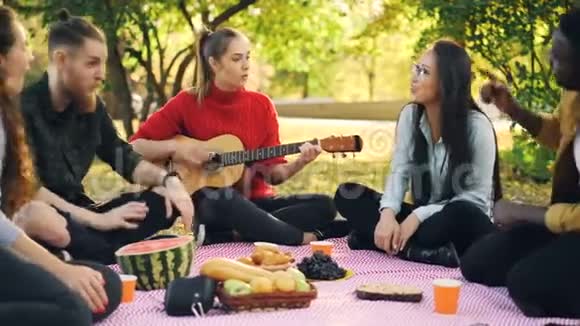 浪漫的年轻人在公园的草坪上坐在格子花格子上野餐一边唱歌一边弹吉他一边玩视频的预览图
