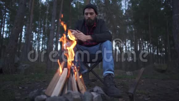 年轻的胡须男坐在椅子上搓着双手取暖森林里的火堆旁视频的预览图
