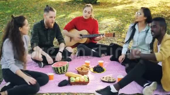 当她的朋友在野餐时听音乐唱歌时一个迷人的女孩在弹吉他视频的预览图