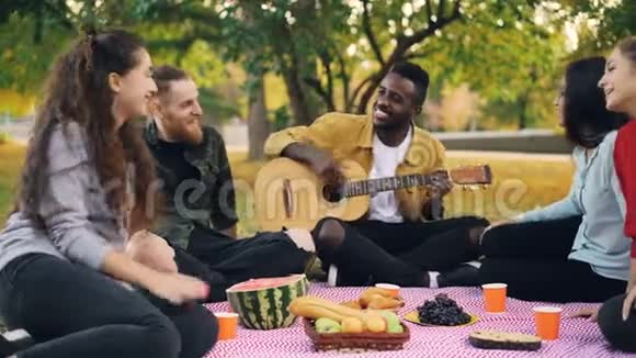 快乐的非裔美国人正在为他的朋友演奏吉他他们坐在野餐的毯子上享受着温暖视频的预览图