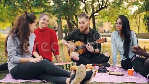 年轻的吉他手在野餐时在户外唱歌和弹吉他而他的朋友们正在听他的音乐视频的预览图