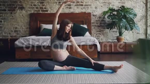 美丽的怀孕女孩正在伸展身体和腿坐在瑜伽垫上用抬起的手向前弯曲不错的阁楼视频的预览图