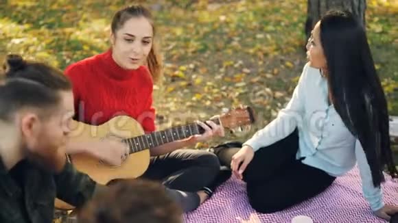 多民族的欢乐朋友正坐在公园的毯子上唱歌漂亮的女人正在演奏视频的预览图