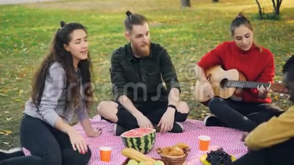 快乐的学生在公园里坐在毯子上享受音乐在野餐时弹吉他唱歌可享用视频的预览图