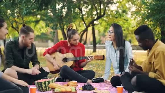 漂亮的女吉他手正在野餐中弹吉他她的朋友们坐在野餐中唱歌拍手视频的预览图