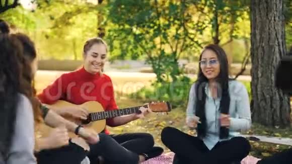 女吉他手正在为坐在城市公园草坪上的快乐朋友弹吉他而男孩和女孩则坐在毯子上视频的预览图