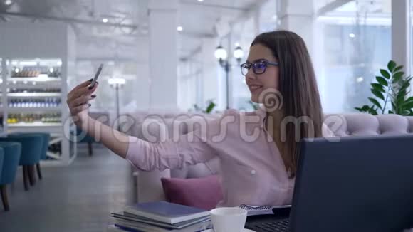 漂亮的餐馆老板女孩在笔记本电脑工作期间用手机拍自拍照片并在笔记本上做笔记视频的预览图