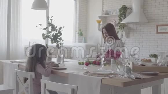 漂亮的年轻微笑的女人站在现代厨房的桌子旁做沙拉坐在椅子上的小女孩视频的预览图