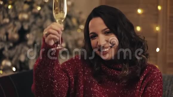 迷人的女人拿着一杯香槟看着圣诞树背景上的相机她举起酒杯视频的预览图