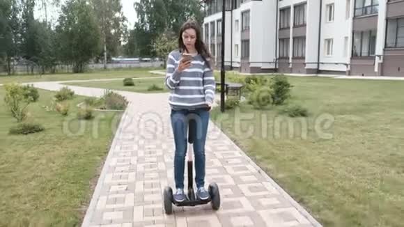 漂亮的黑发女人在家附近骑着陀螺滑板车看着手机视频的预览图