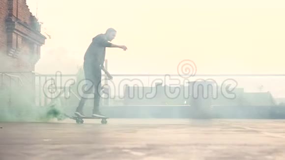 英俊的年轻人滑板骑着绿色的烟雾在工业建筑的露台上滑板运动员在做视频的预览图
