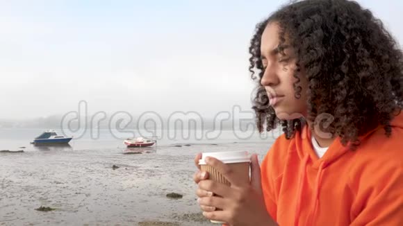 穿着橙色连帽衫的年轻的混血女人在港口边喝着外卖咖啡看上去悲伤或体贴视频的预览图