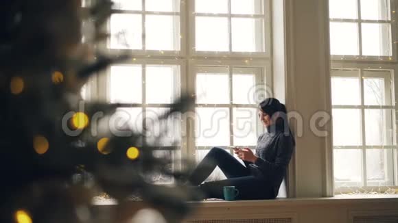 快乐的年轻女性使用智能手机坐在窗台上度过圣诞节在家放松享受现代生活视频的预览图