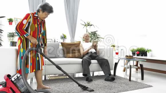 快乐的亚洲资深男子在家玩平板电脑和资深女子吸尘地板视频的预览图