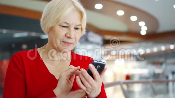 商场的高级妇女使用手机在购物中心浏览手机的女购物者视频的预览图