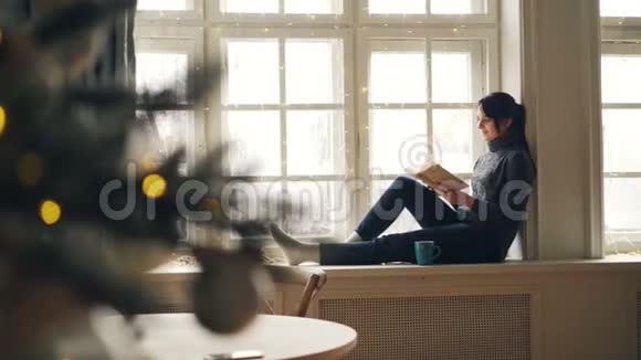微笑着的年轻女子正在阅读圣诞节那天坐在窗台上的书享受休息和平静的假期视频的预览图