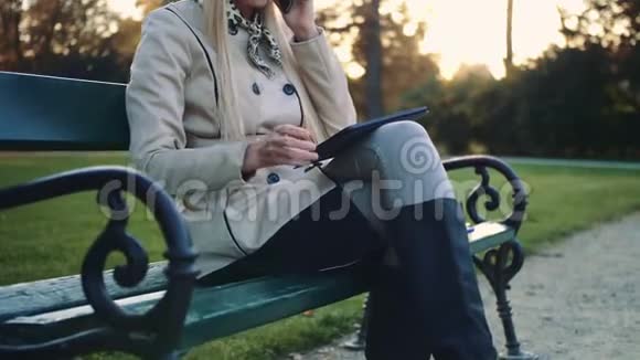 漂亮的女孩坐在长凳上平板电脑和手机坐在公园的长凳上视频的预览图