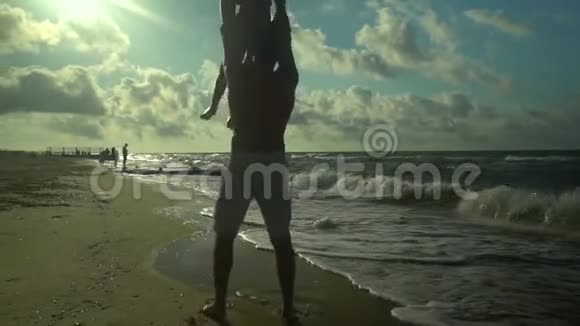 一位年轻的父亲和他的儿子沿着海滨散步阳光灿烂爸爸把他的儿子旋转把他扔进水里视频的预览图