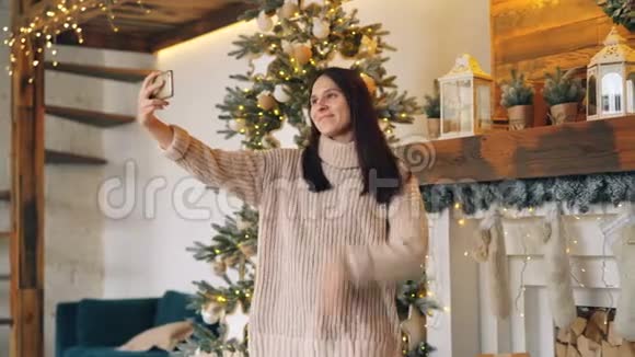 圣诞节那天美丽的女孩正站在壁炉和冷杉旁用智能手机进行在线视频通话视频的预览图