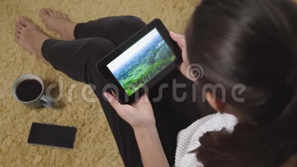 女人在地毯上坐在家里用平板电脑在屏幕上观看大自然和度假视频视频的预览图