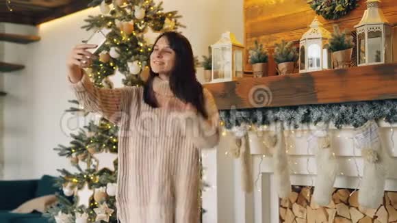圣诞节当天一位无忧无虑的女孩正站在圣诞树旁壁炉旁拿着智能手机摆姿势自拍视频的预览图