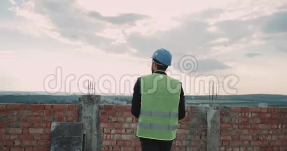 年轻的工程师带着安全帽和高能见度背心从后面拍屋顶建筑背景的照片视频的预览图