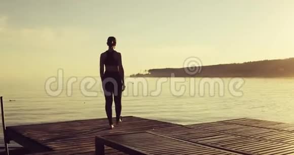 日落时分女人在河边的码头上用树姿练习瑜伽后景视频的预览图
